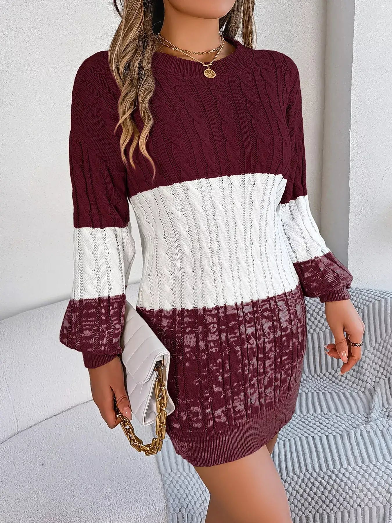Woman wearing a color block cable knit sweater dress with textured pattern, featuring maroon and white sections, holding a clutch.