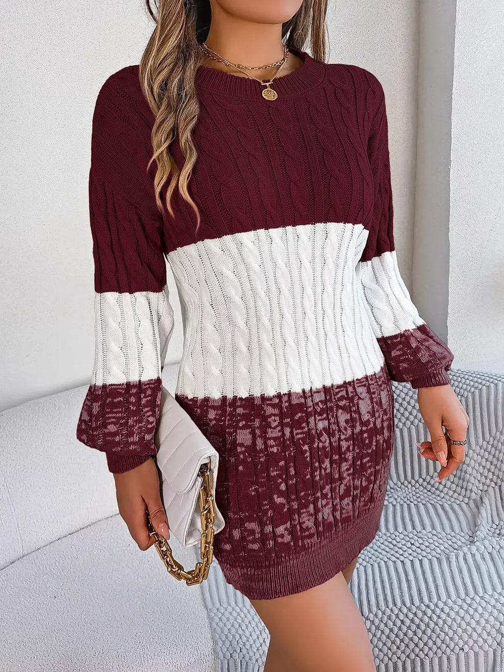 Woman wearing a color block cable knit sweater dress with textured pattern, featuring maroon and white sections, holding a clutch.