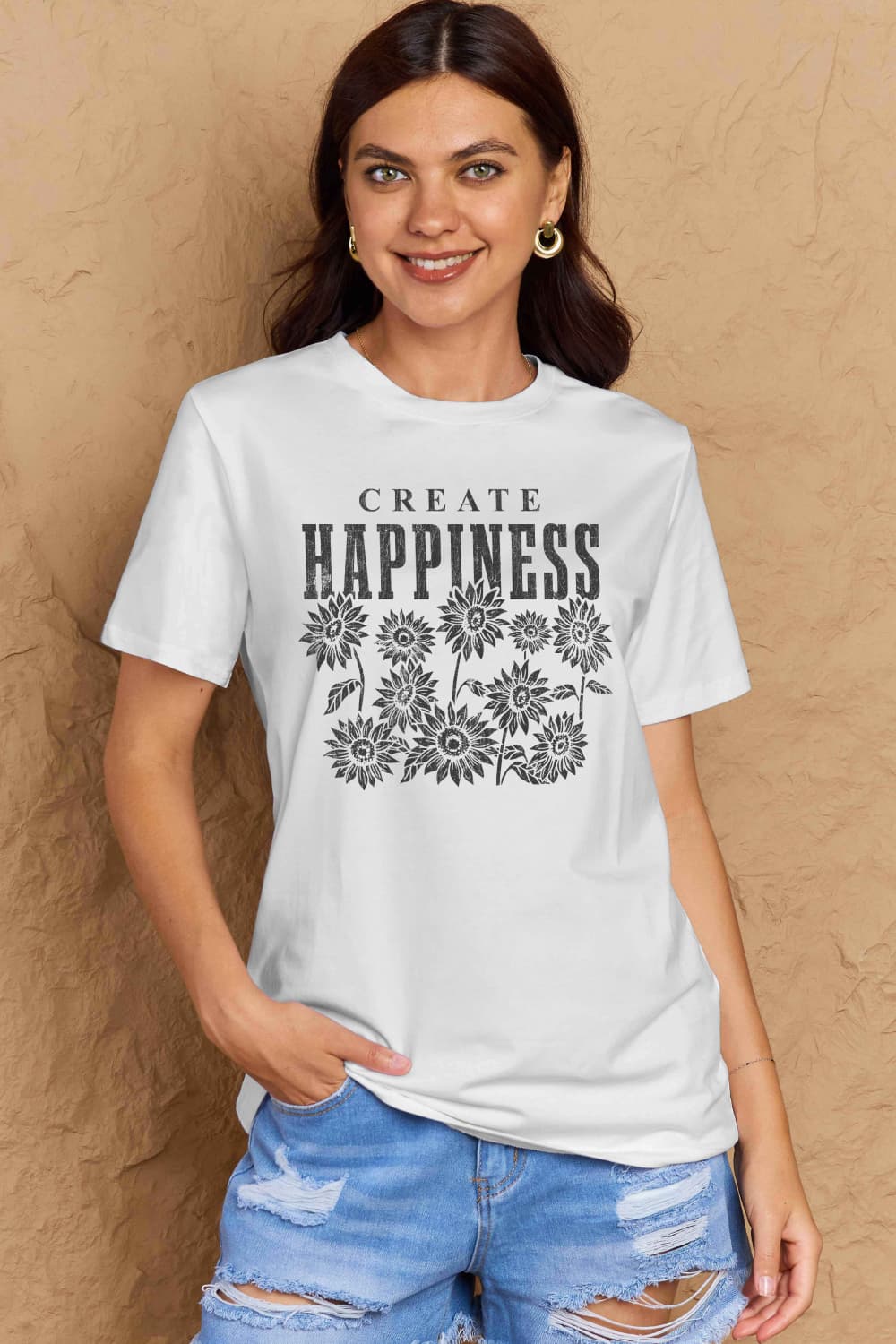 Woman wearing a Graphic Cotton T-Shirt with 'CREATE HAPPINESS' print, styled casually with ripped jeans, promoting positivity.